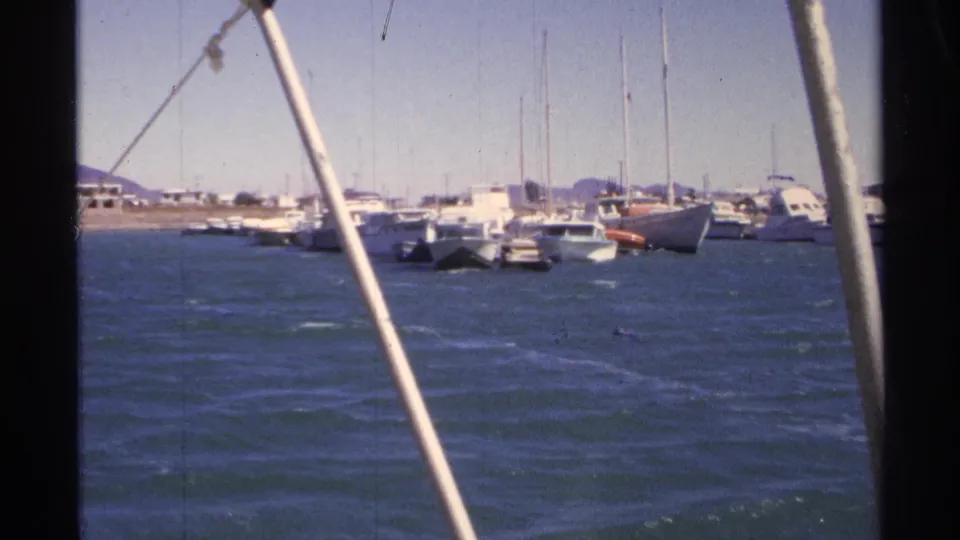 1975 Guaymas harbor view through foreground rigging, with choppy water, moored sailboats, small craft, and distant shore.