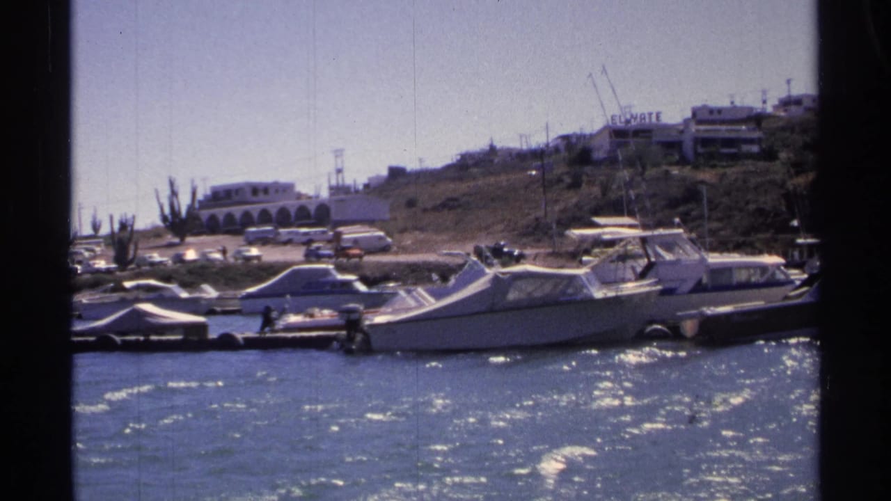 Motorboats docked by shore with cars, hillside buildings, and an El Yate sign behind them.