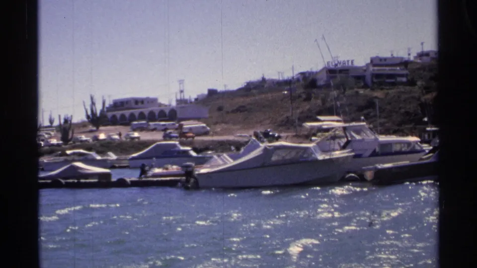 Docked boats along the Guaymas waterfront with parked cars, hillside structures, distant people, and a visible El Yate sign.