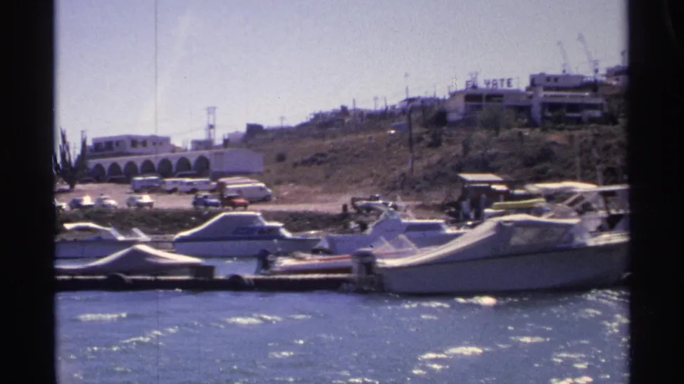 Guaymas marina boats seen from the water, with parked cars, small figures, hillside buildings, and the El Yate sign in the background.