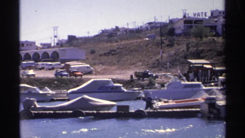 Late-clip marina view in Guaymas with small boats, a floating dock, shoreline vehicles, hillside buildings, and the El Yate sign.