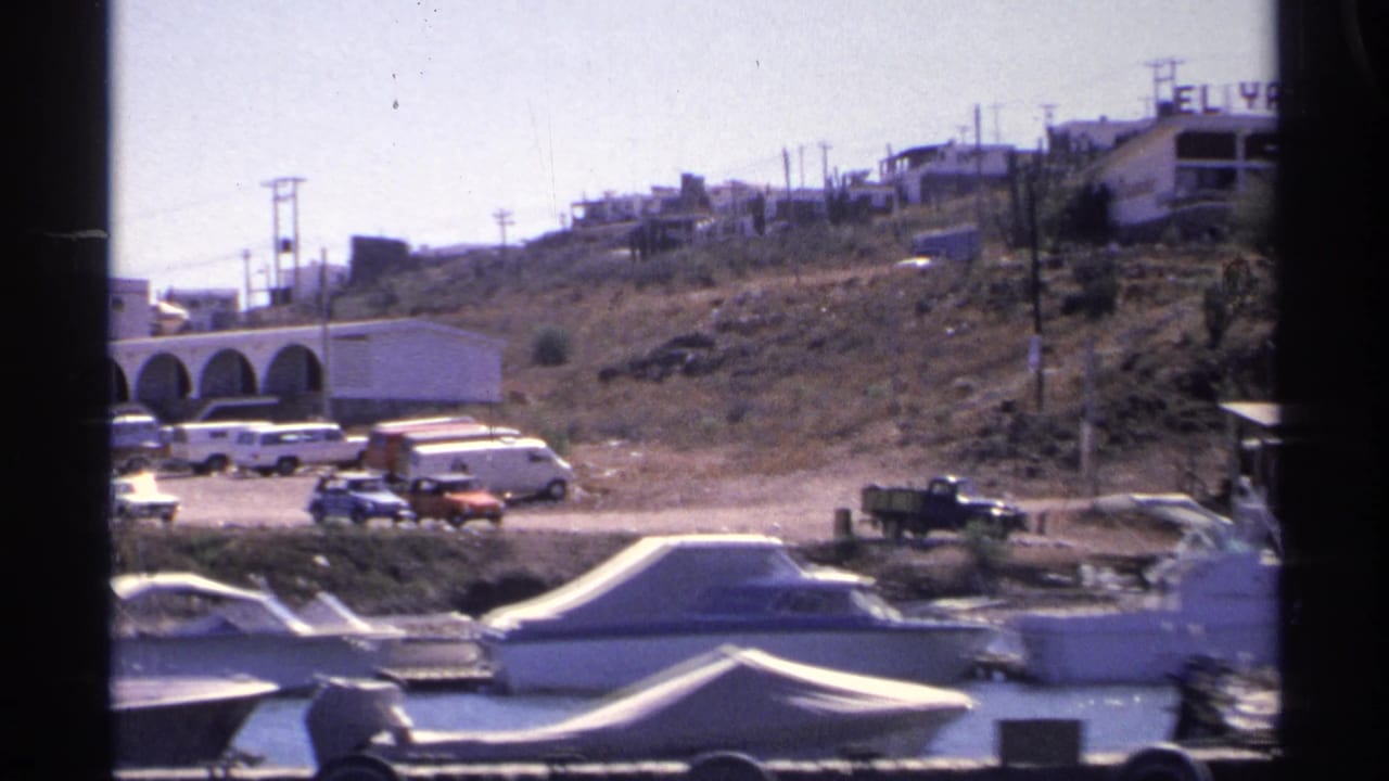 1975 Guaymas harbor view with boats in front, 1970s vehicles on a dirt lot, hillside buildings, utility poles, and a distant EL YATE sign.