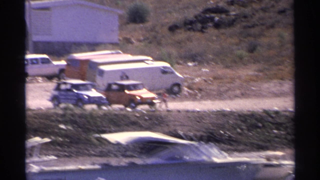 1975 Guaymas harbor parking area with white and orange vans, compact cars, a white building, dry hillside, and boat roofs near the water.