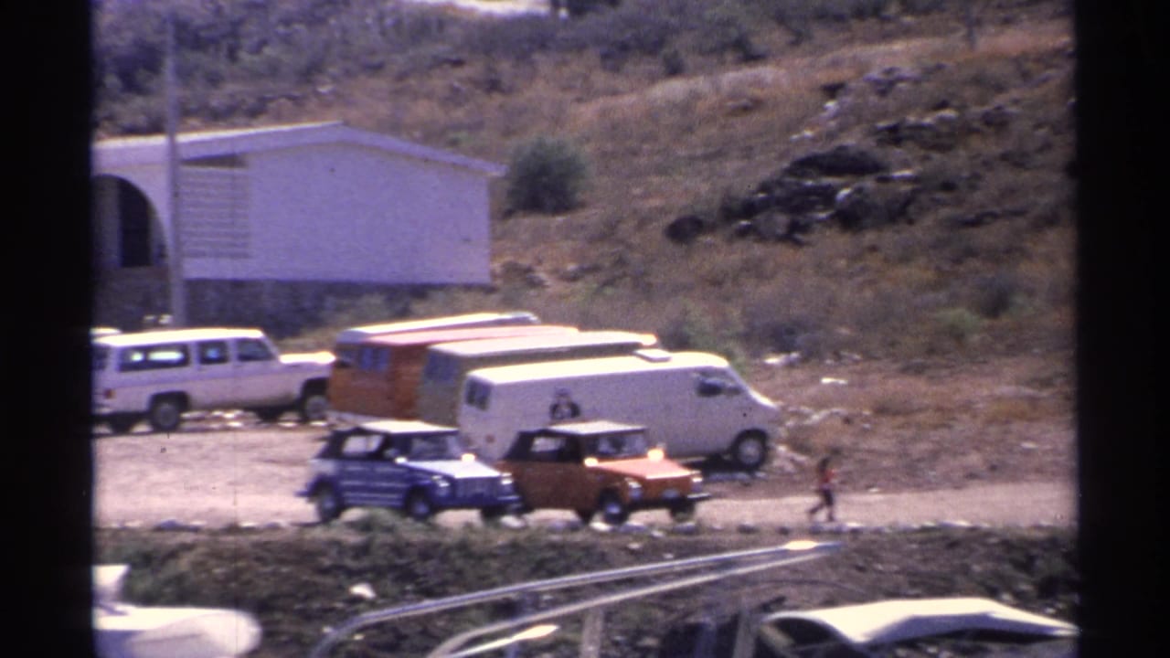 Guaymas harbor frame with 1970s vehicles parked on a dirt lot, boats along the lower edge, a white arched building, and a distant person walking.