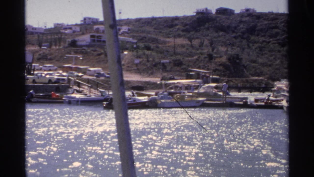 1975 Guaymas marina view across bright water, with moored small boats, people on the dock, parked cars, and hillside buildings beyond.