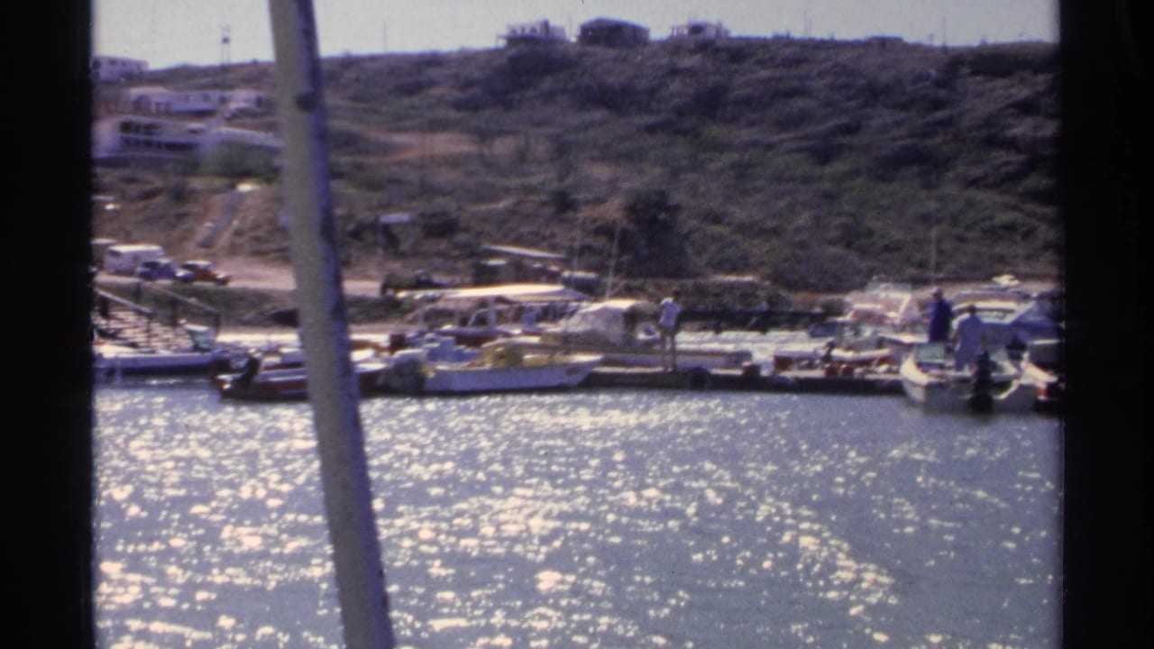 A 1975 Guaymas dock scene shows small craft tied along the marina, distant people, parked vehicles, and dry hillside terrain behind.