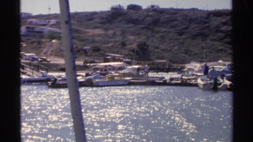 A 1975 Guaymas dock scene shows small craft tied along the marina, distant people, parked vehicles, and dry hillside terrain behind.