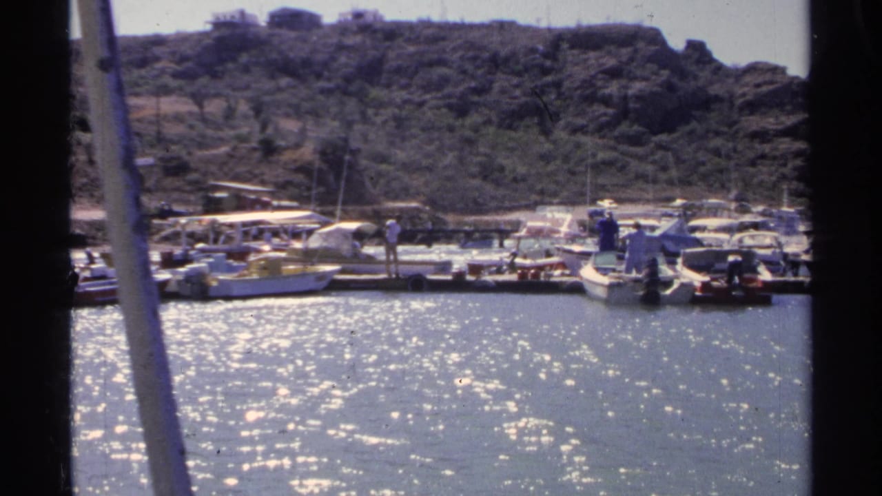 Guaymas marina view with bright reflected water, people near moored boats, dock structures, and a scrub-covered hillside in the distance.