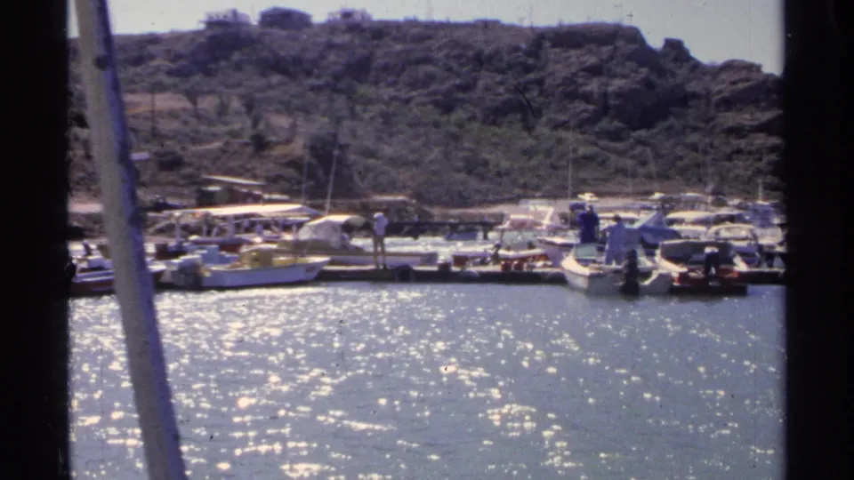 Guaymas marina view with bright reflected water, people near moored boats, dock structures, and a scrub-covered hillside in the distance.