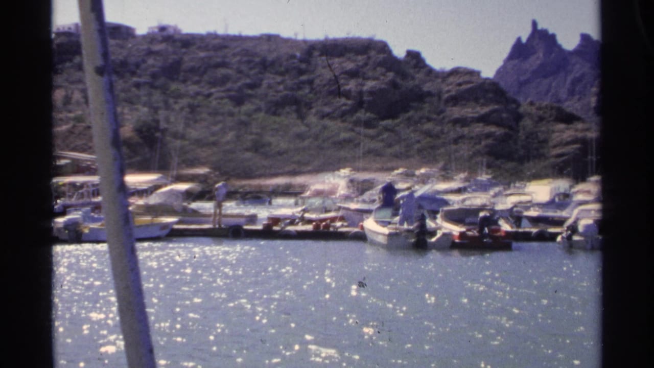 Final 1975 Guaymas marina frame shows clustered small boats, distant dockside figures, glittering water, and jagged hillside rocks beyond.