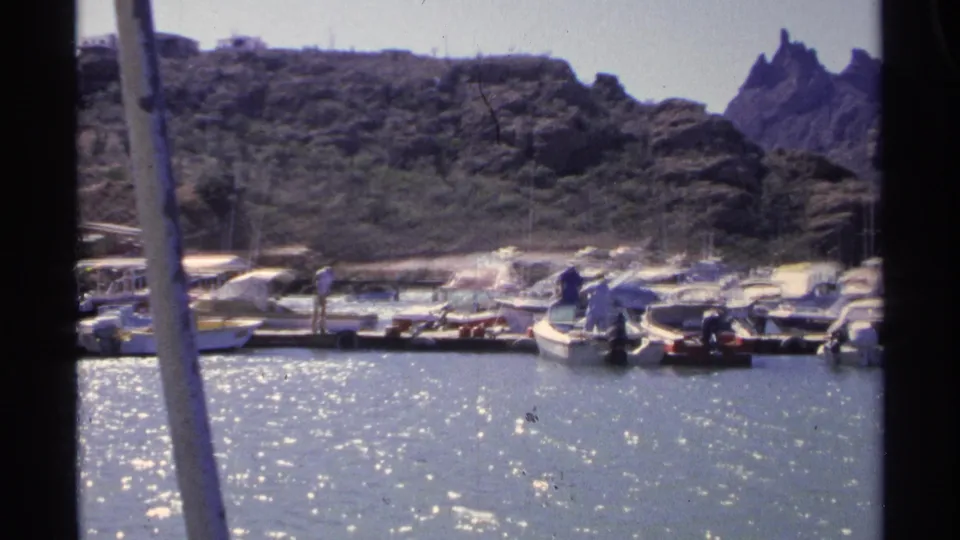 Final 1975 Guaymas marina frame shows clustered small boats, distant dockside figures, glittering water, and jagged hillside rocks beyond.