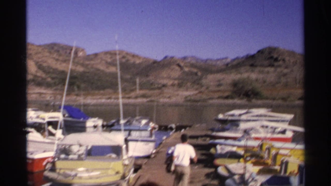 1975 Guaymas, Mexico marina view with small boats along a dock, sailboat masts, calm water, and arid hills beyond.
