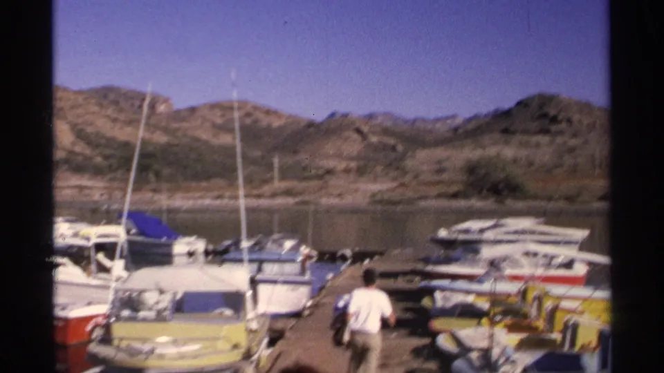 1975 Guaymas, Mexico marina view with small boats along a dock, sailboat masts, calm water, and arid hills beyond.