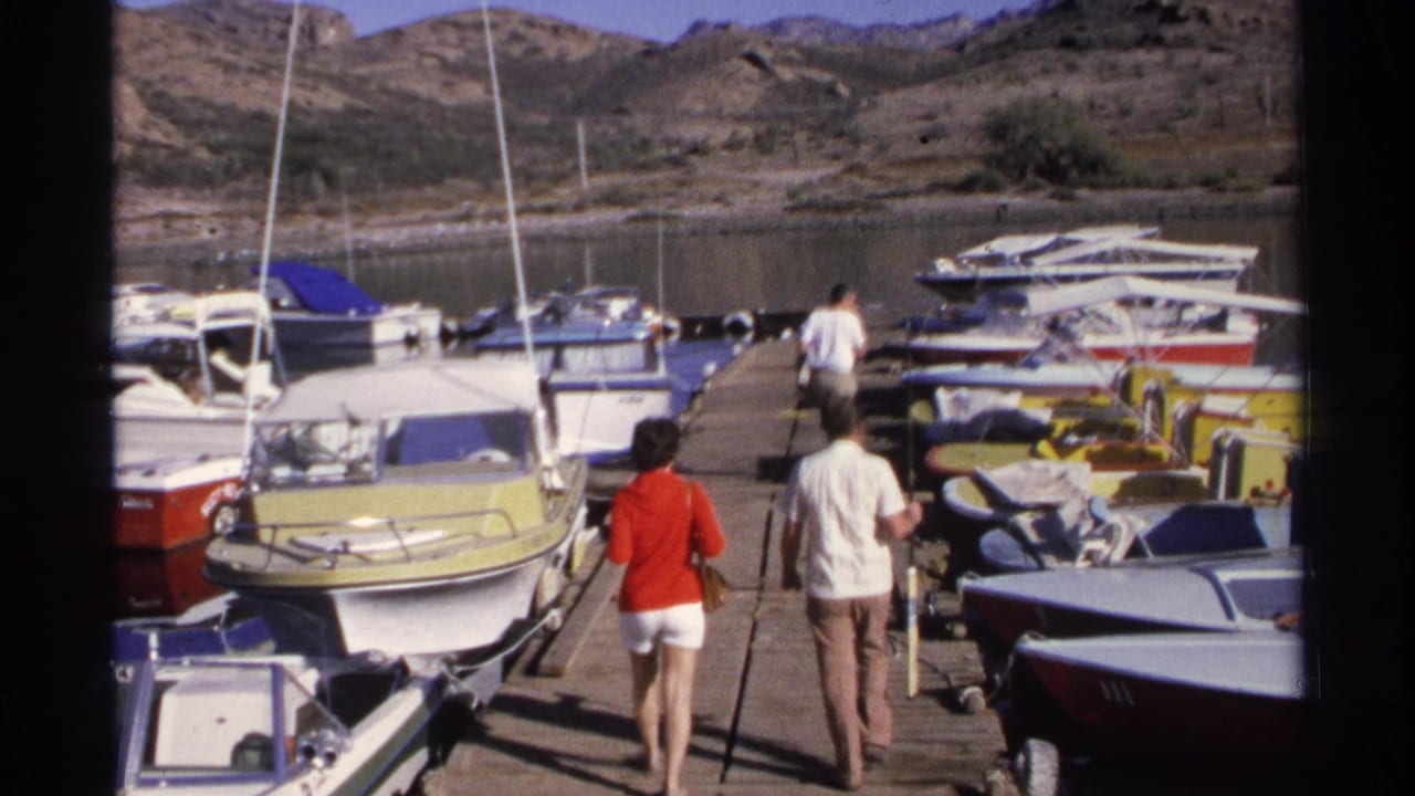 A woman and two men walk along a Guaymas marina dock between small boats, with masts, water, and dry hills behind them.