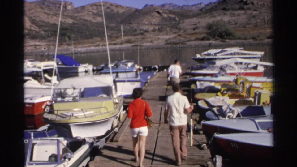 A woman and two men walk along a Guaymas marina dock between small boats, with masts, water, and dry hills behind them.