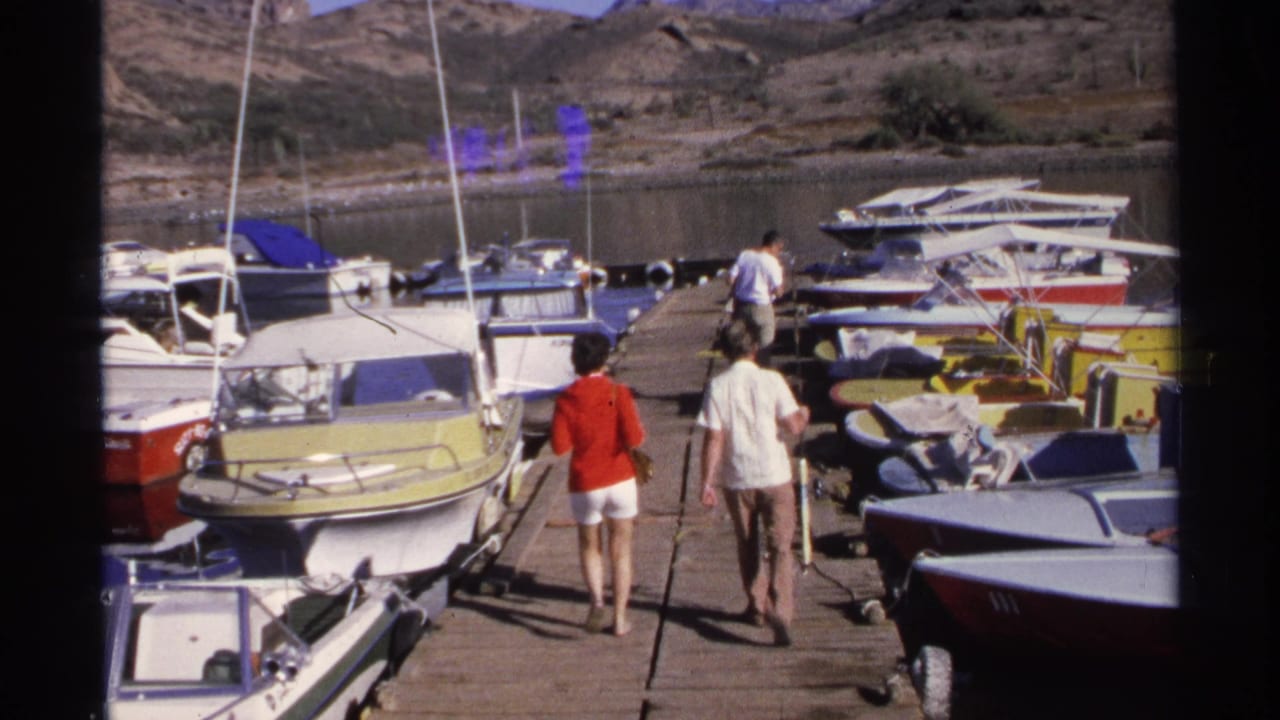 1975 Guaymas home-movie frame showing two adults walking along a wooden pier lined with small motorboats under bright sun.