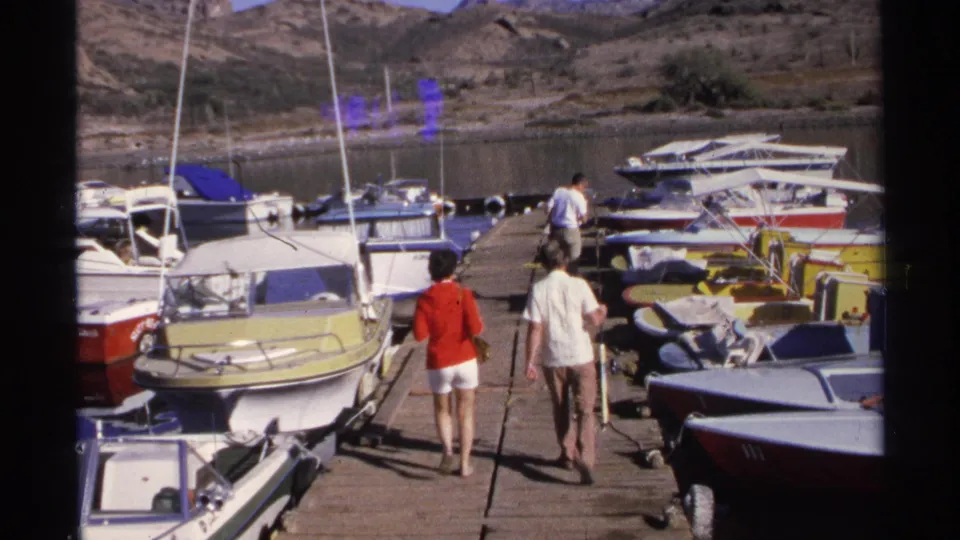 1975 Guaymas home-movie frame showing two adults walking along a wooden pier lined with small motorboats under bright sun.