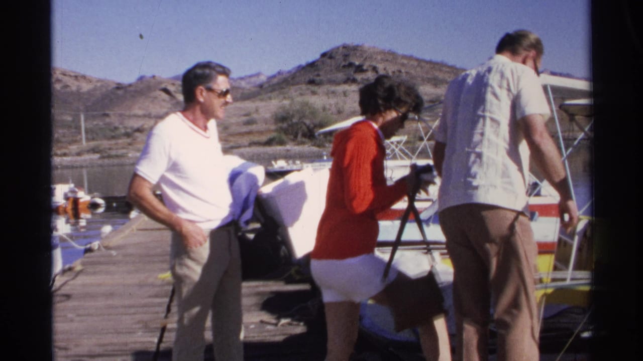 1975 Guaymas home-movie frame showing three adults near boats on a wooden pier, with arid hills and water behind them.