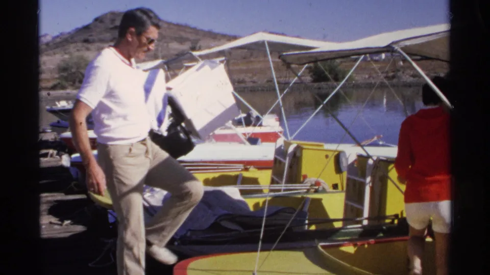 In 1975 Guaymas, Mexico, a man carrying a white cooler steps toward a yellow and white boat as a woman in red stands nearby.