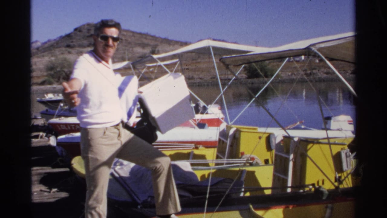 A man in sunglasses carries a white cooler beside a small boat at a Guaymas dock, with water and low hills behind him.