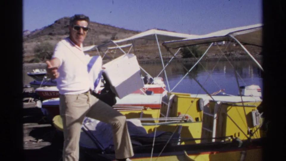 A man in sunglasses carries a white cooler beside a small boat at a Guaymas dock, with water and low hills behind him.