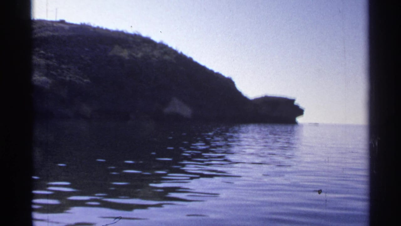 1975 Guaymas home-movie frame showing calm water, a dark rocky slope, and pale sky viewed through a vertical frame edge.