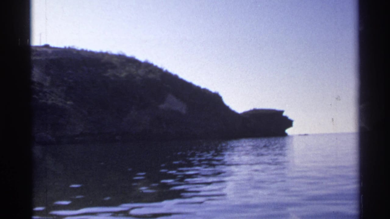 1975 Guaymas frame with a low rocky hill, calm reflective water, pale sky, and dark vertical borders from the filming position.