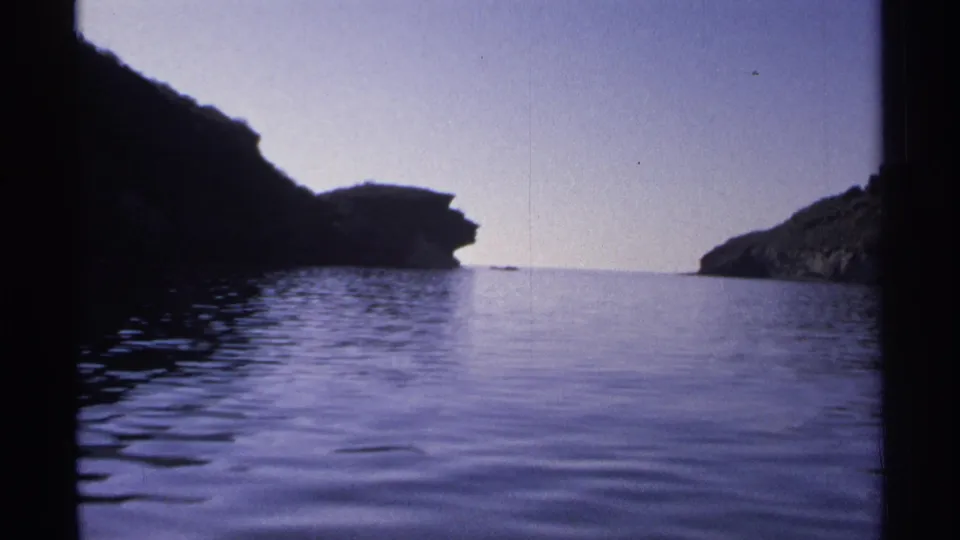 1975 Guaymas home-movie frame showing a boat-level view across calm water toward dark coastal cliffs and open sky.