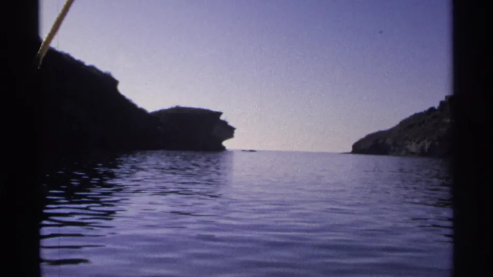 1975 Guaymas frame from a moving boat, with dark cliff edges, still blue water, and a rocky coastal opening ahead.