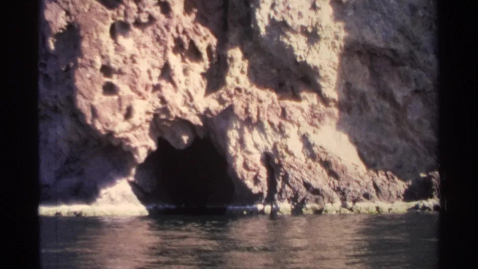 1975 Guaymas home-movie frame showing a sunlit cliff face, a dark cave opening, and reflective water at the shoreline.