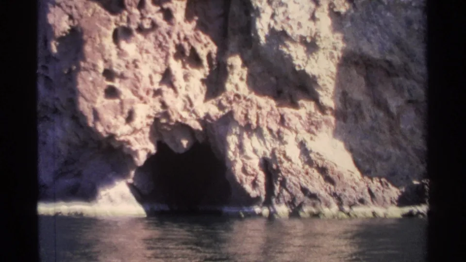 1975 Guaymas frame with rough sunlit stone, a shadowed cave entrance, and calm water viewed from a boat.