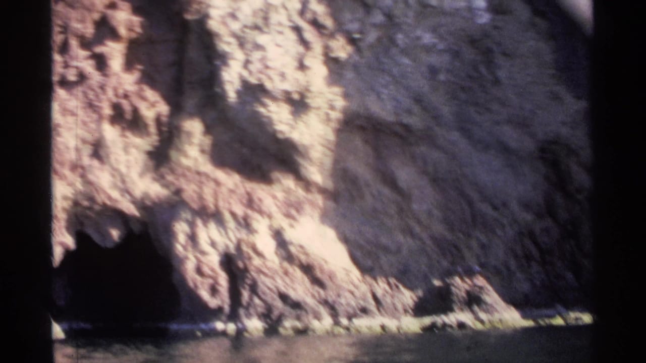 Rocky cliff face with a dark cave opening at the waterline and calm water below.