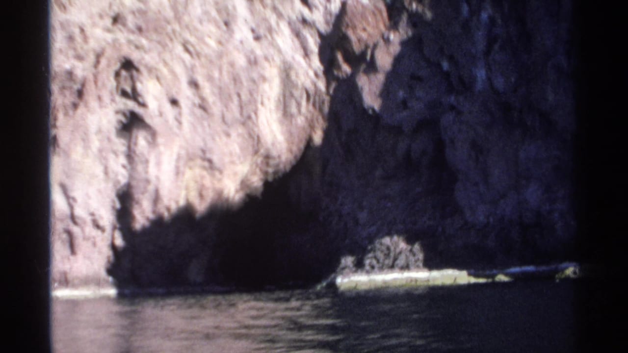 1975 Guaymas 8mm frame with a steep coastal rock wall, heavy shadow, and still water below.