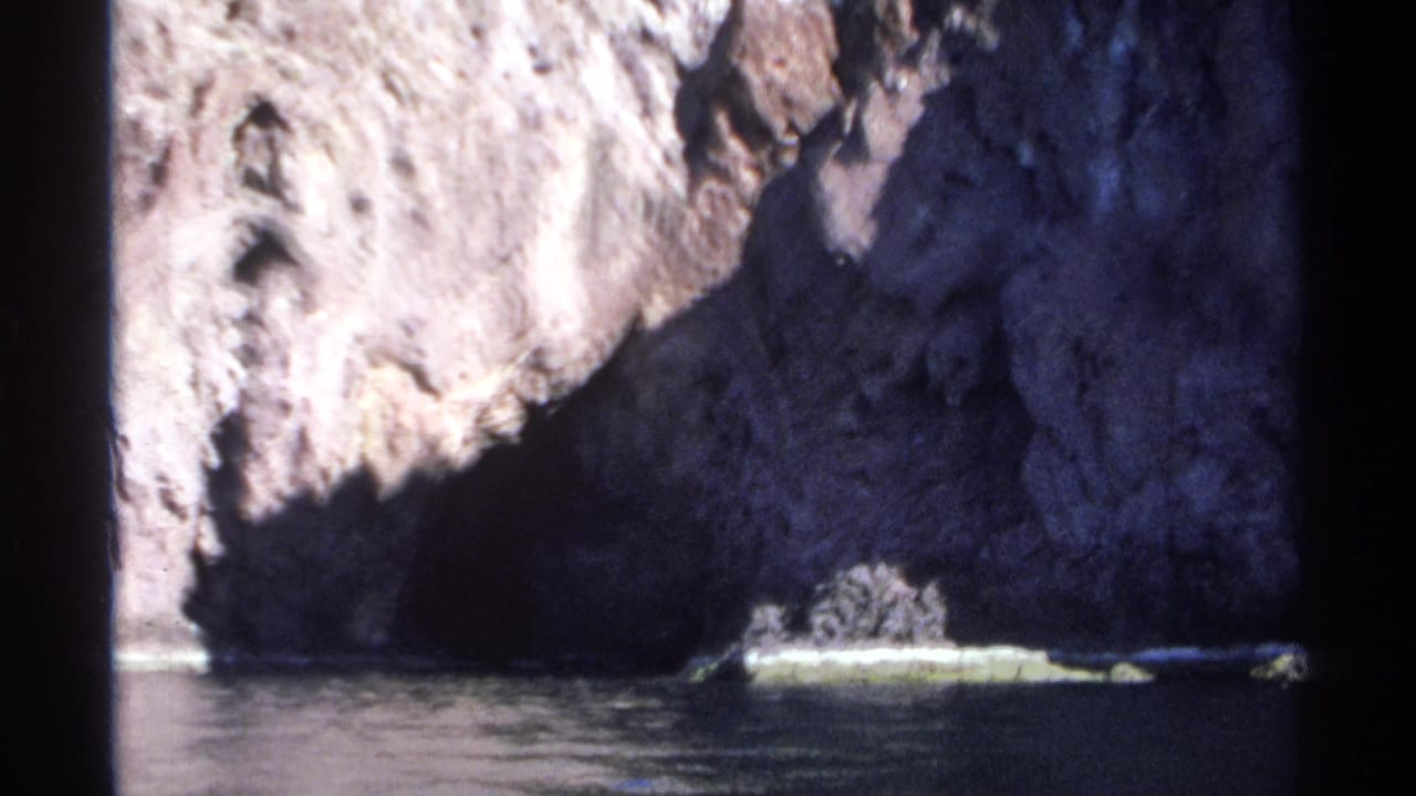 Rocky cliff and dark cave entrance at the waterline seen from a boat.