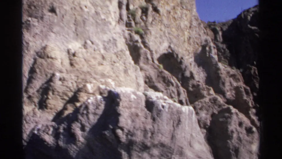 1975 Guaymas travel frame showing a textured canyon wall, small patches of vegetation, and blue sky from a low river-level perspective.