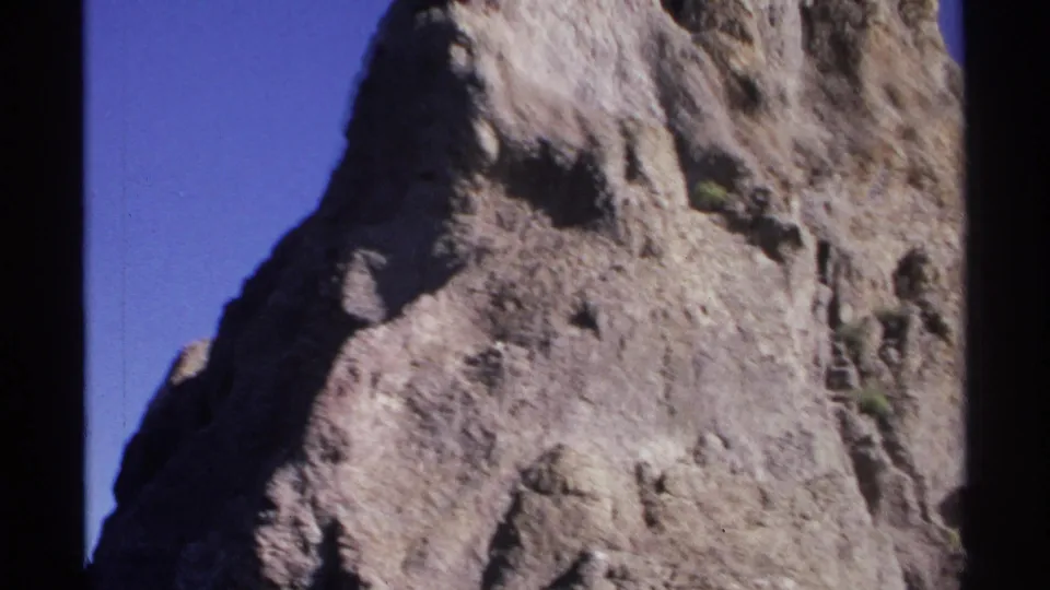1975 Guaymas canyon footage ending on a high, textured rock face rising against clear blue sky, framed by dark 8mm gate edges.