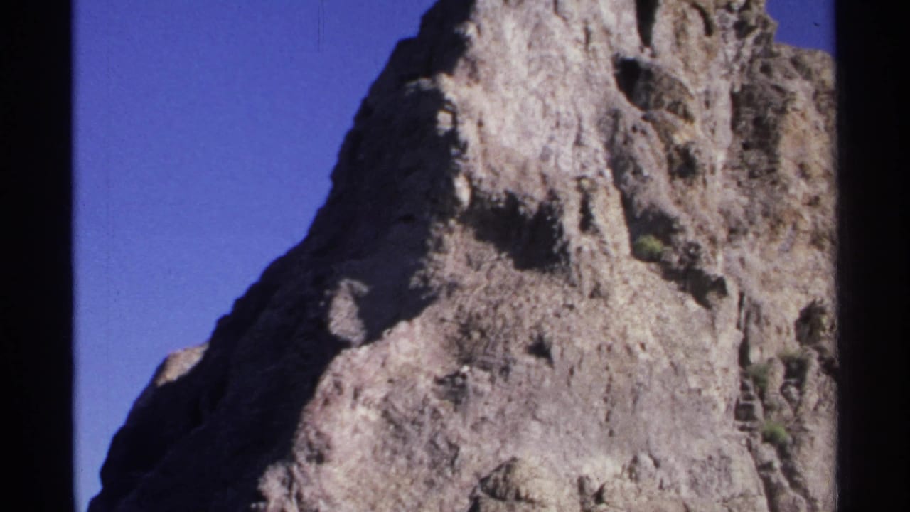 Steep gray rock face rising against a clear blue sky with black film-frame edges.