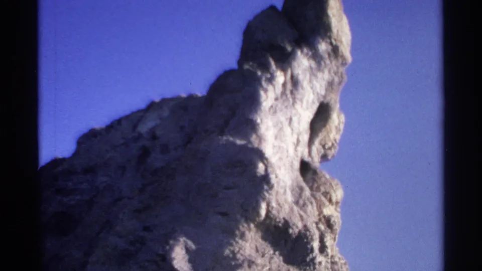 1975 Guaymas 8mm frame showing a craggy sunlit rock peak, dark cavities, and deep shadows beneath a clear sky.