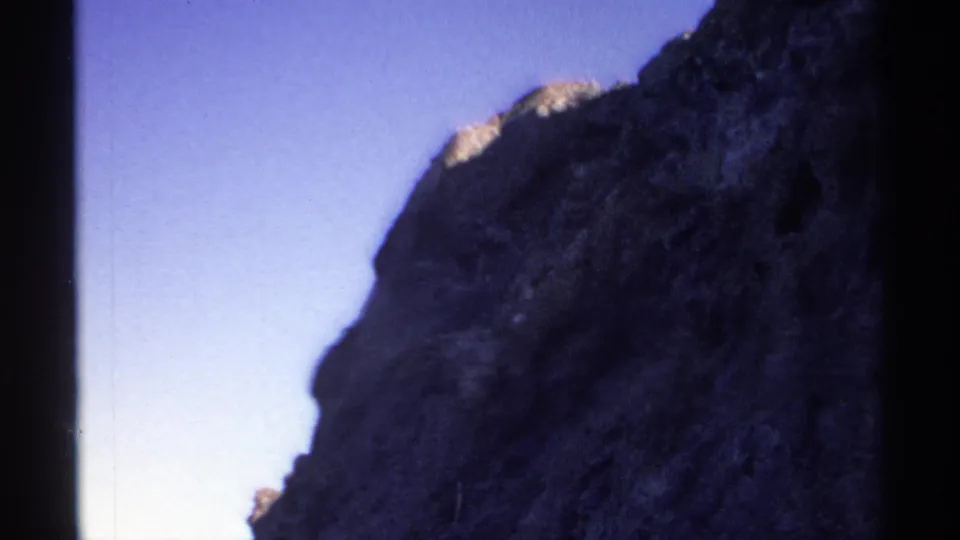 1975 8mm Guaymas frame showing a close, shadowed cliff edge against clear blue sky with visible film grain.