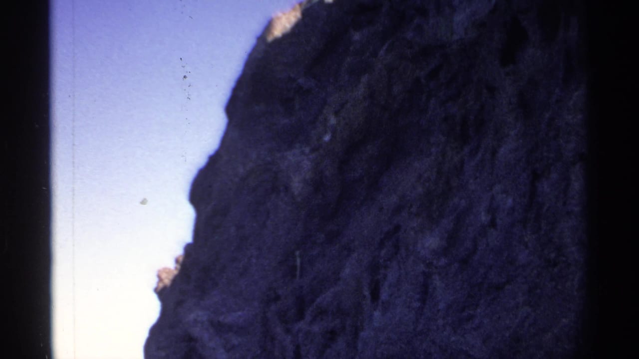 Dark rocky cliff beside blue water under a pale sky.
