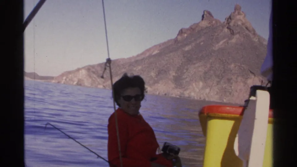 A smiling woman in a red jacket and sunglasses sits aboard a small boat off Guaymas with rocky mountains behind her.