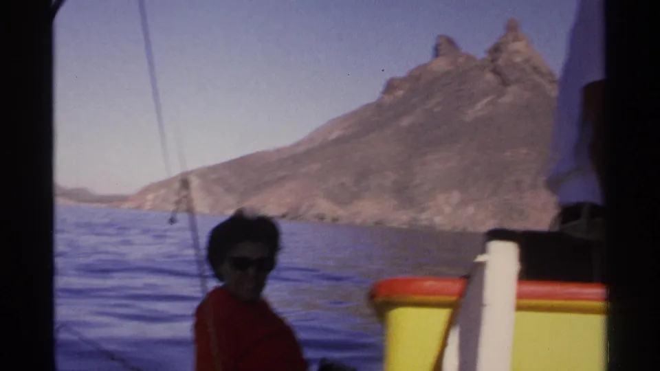 A woman aboard a small boat is seen against calm water and arid mountains in Guaymas, Mexico, on 1970s 8mm footage.