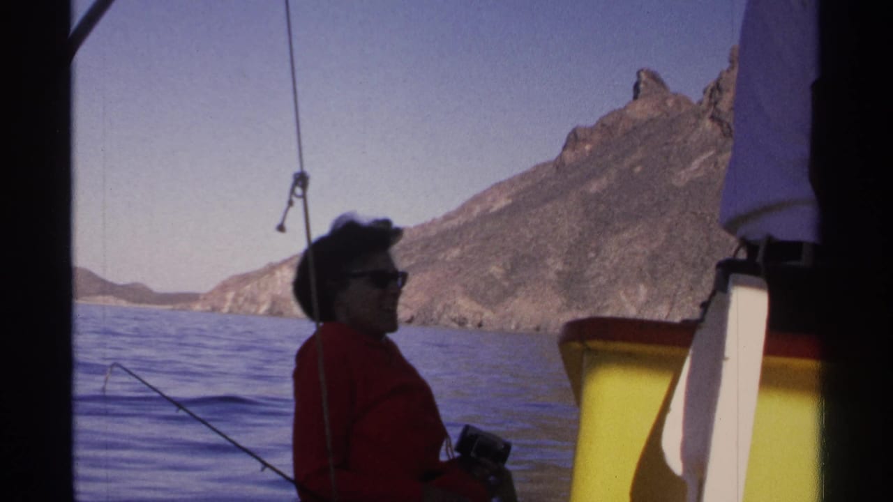 1975 Guaymas home-movie frame showing a woman in a red top on a boat, holding a fishing rod beside blue water and a rugged coastal mountain.
