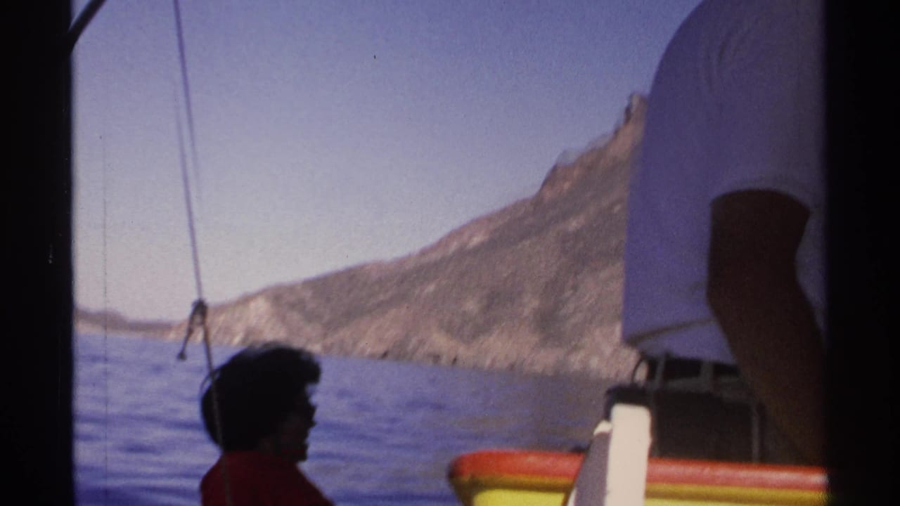 1975 Guaymas boat scene with the seated woman partly in shadow, a man in a white shirt at right, blue water, rigging, and mountain shoreline.