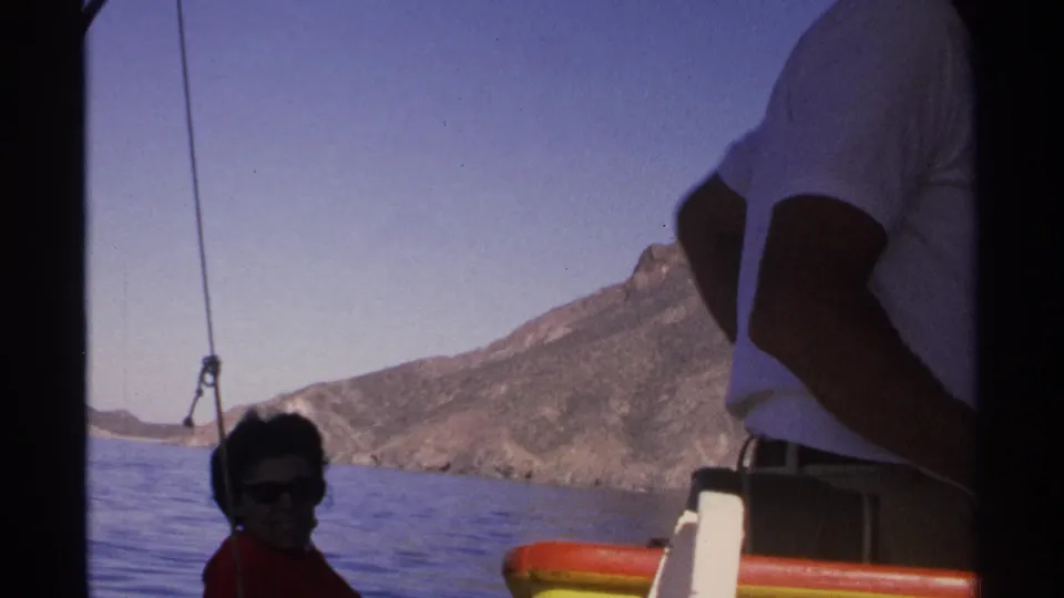 1975 Guaymas home-movie view from a boat, with the woman seated in shadow, the man standing nearby, and a rugged mountain across the water.
