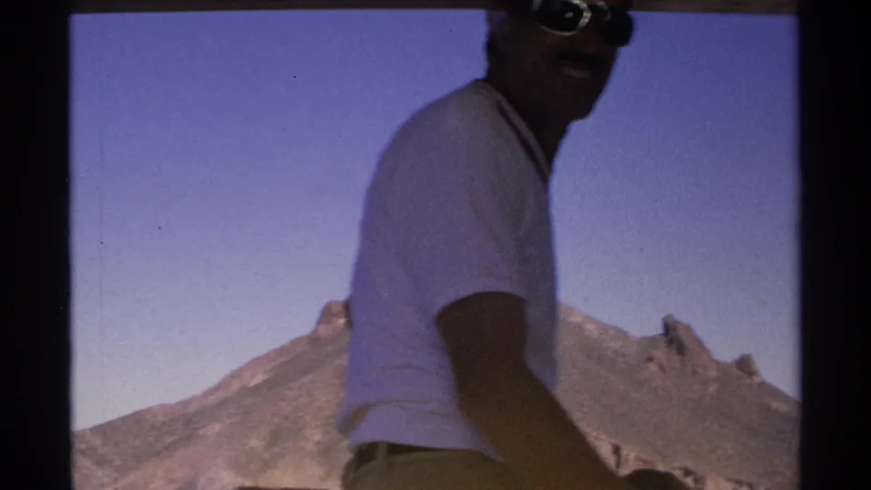 1975 Guaymas frame showing a man in a white shirt and sunglasses smiling on the boat, with clear sky and coastal mountain terrain behind him.