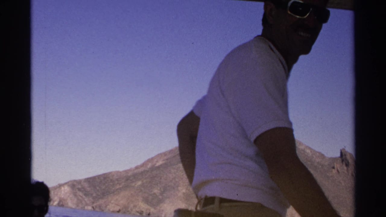1975 8mm home-movie frame from Guaymas showing a man in sunglasses on a small boat with rugged mountains beyond the water.