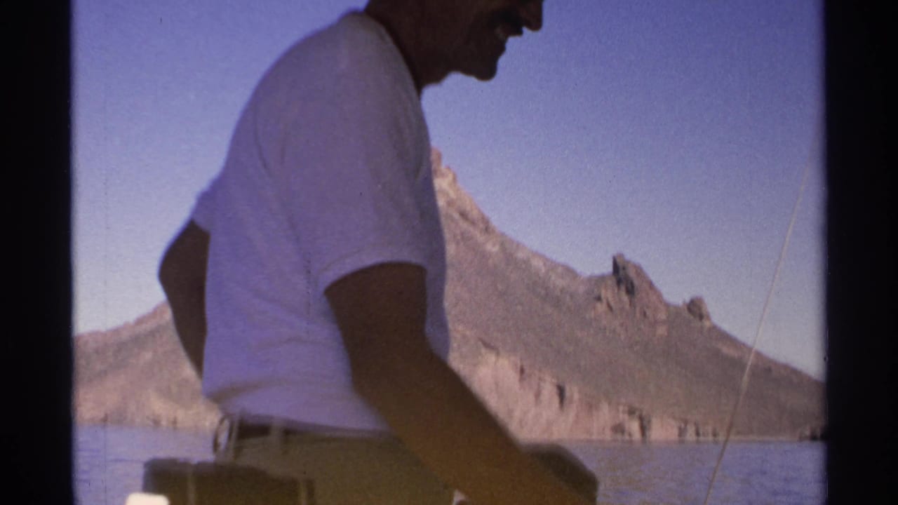 Man in a white shirt standing on a boat with mountains across the water.