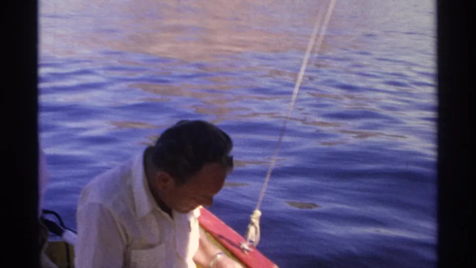 1975 8mm frame from Guaymas showing a seated man focused downward beside a fishing line on calm blue water.