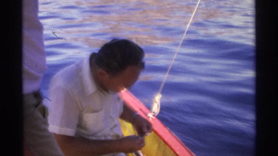 1975 Guaymas home-movie frame showing a man seated by a red boat rail, looking down as he works with a fishing line.